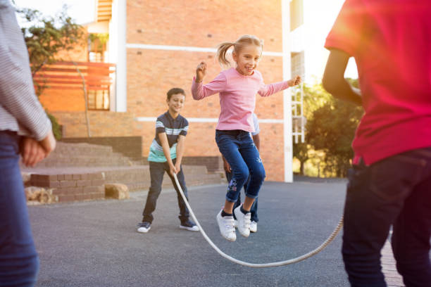 ing rope outdoor. Children playing skipping rope jumping game and laughing outdoors. Happy cute girl jumping over skipping rope held by her friends.